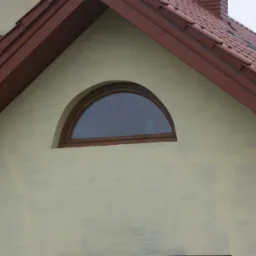 Półokrągłe okno z brązową ramą na szczycie budynku z beżową elewacją i czerwoną dachówką. Ujęcie z dołu, lekko z prawej strony.