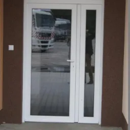Białe, przeszklone drzwi wejściowe z nadświetlem, zamontowane w brązowej elewacji. W odbiciu szyby widoczny samochód ciężarowy i osoba.