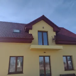 Żółty dom jednorodzinny z nowym, bordowym dachem z dachówki ceramicznej, bordowymi rynnami i oknami z brązowymi ramami, widziany z dołu w słoneczny dzień.