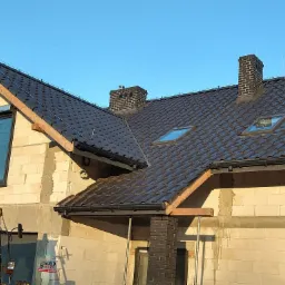 Dom w trakcie budowy z częściowo ukończonym ciemnobrązowym dachem z dachówki ceramicznej, widoczne okna dachowe, komin z cegły klinkierowej, orynnowanie oraz żółta koparka z przodu budynku.