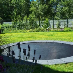 Zadbany trawnik z wbudowaną trampoliną otoczoną szarą obudową, w tle osoba pracująca w ogrodzie przy siatce ogrodzeniowej, z przodu widoczne kwitnące lawendy.