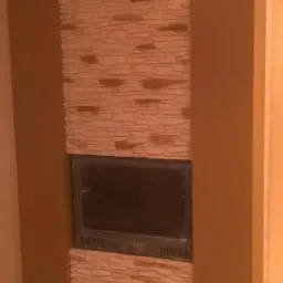 Wykończony kominek z obudową z kamienia dekoracyjnego, otoczony brązowymi panelami, w jasnym wnętrzu. Widoczne detale wykończenia i struktura kamienia.