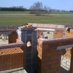 Wznoszenie ścian domu z czerwonej cegły ceramicznej. Widoczny komin, nadproża i wieniec. W tle krajobraz polny i budowa w toku. Prace murarskie w słoneczny dzień.