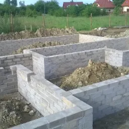 Wznoszenie fundamentów z bloczków betonowych na działce. Widoczne ściany fundamentowe, ziemia i trawa w tle. Budowa w toku, z dbałością o detale.