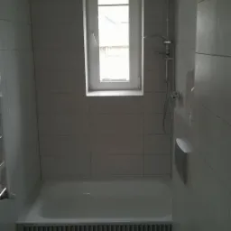 Nowoczesna łazienka w szarościach z wanną, toaletą i prysznicem, wykończona podłużnymi płytkami ceramicznymi, z oknem w białej ramie w tle.