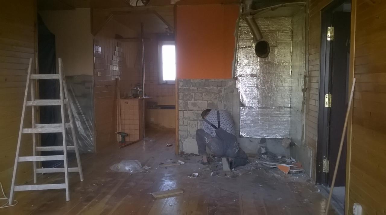 Remont wnętrza domu: pracownik w trakcie skuwania tynku przy ścianie z cegieł, widoczne narzędzia, drabina i fragmenty izolacji. W tle okno i fragment pomieszczenia.