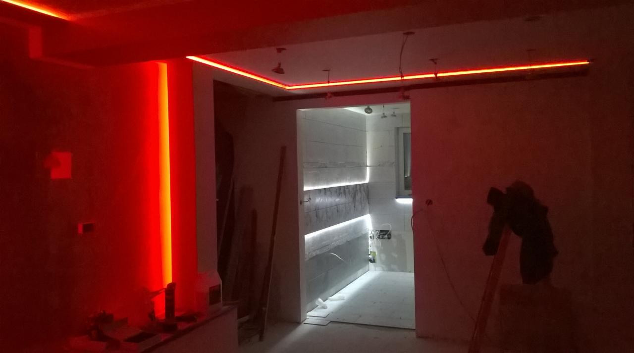 Nowoczesne oświetlenie LED w surowym wnętrzu: czerwone listwy na ścianach i suficie, białe w łazience. Widoczne drabina i narzędzia sugerują etap wykończeniowy.