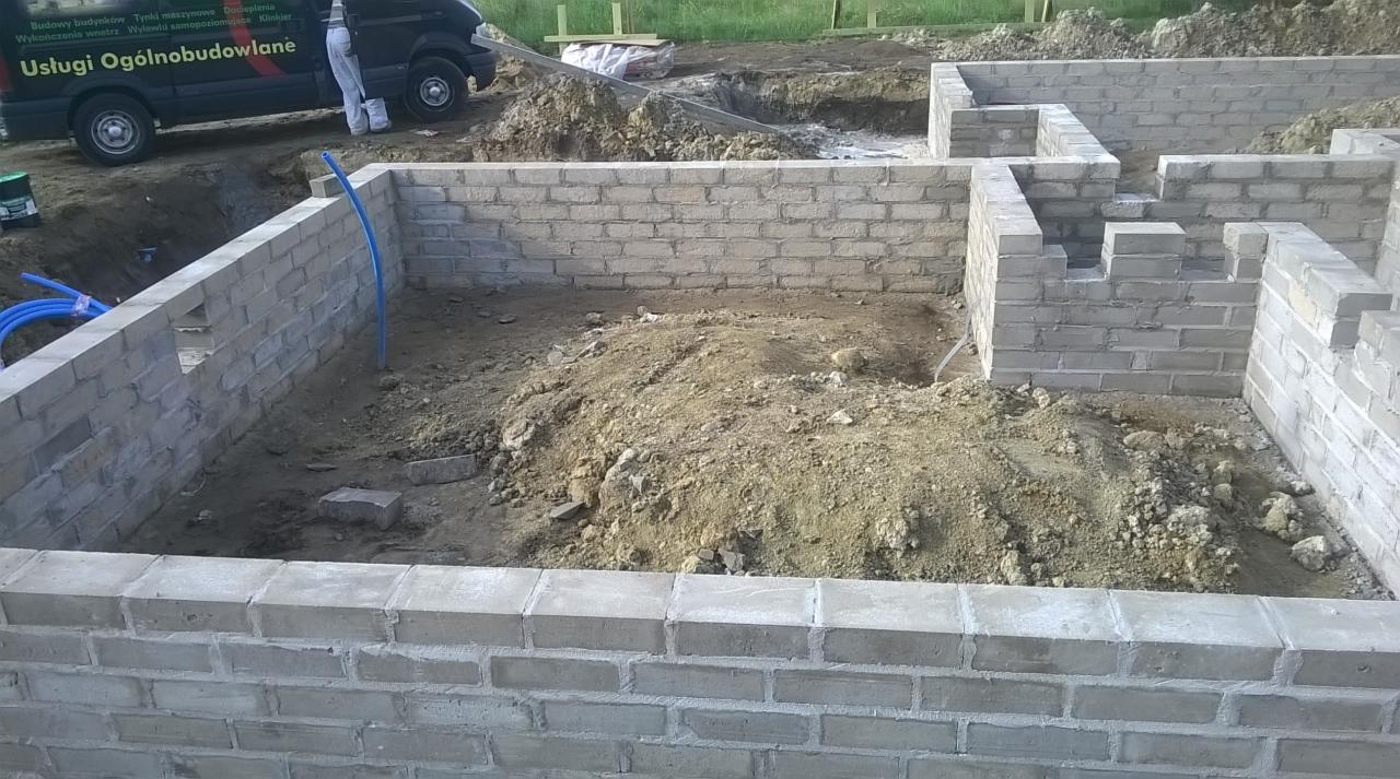 Wymurowane ściany fundamentowe z pustaków szalunkowych, widoczne instalacje i wykop, w tle samochód dostawczy z reklamą usług budowlanych.