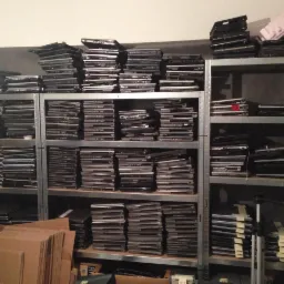 Regał magazynowy wypełniony stertami używanych laptopów, przygotowanych do naprawy lub recyklingu. W tle ściana, w dolnej części kadru kartony.