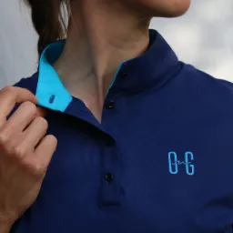 Kobieta w granatowej koszulce polo z turkusowym kołnierzykiem i wyhaftowanym logo, rozpinająca górny guzik.