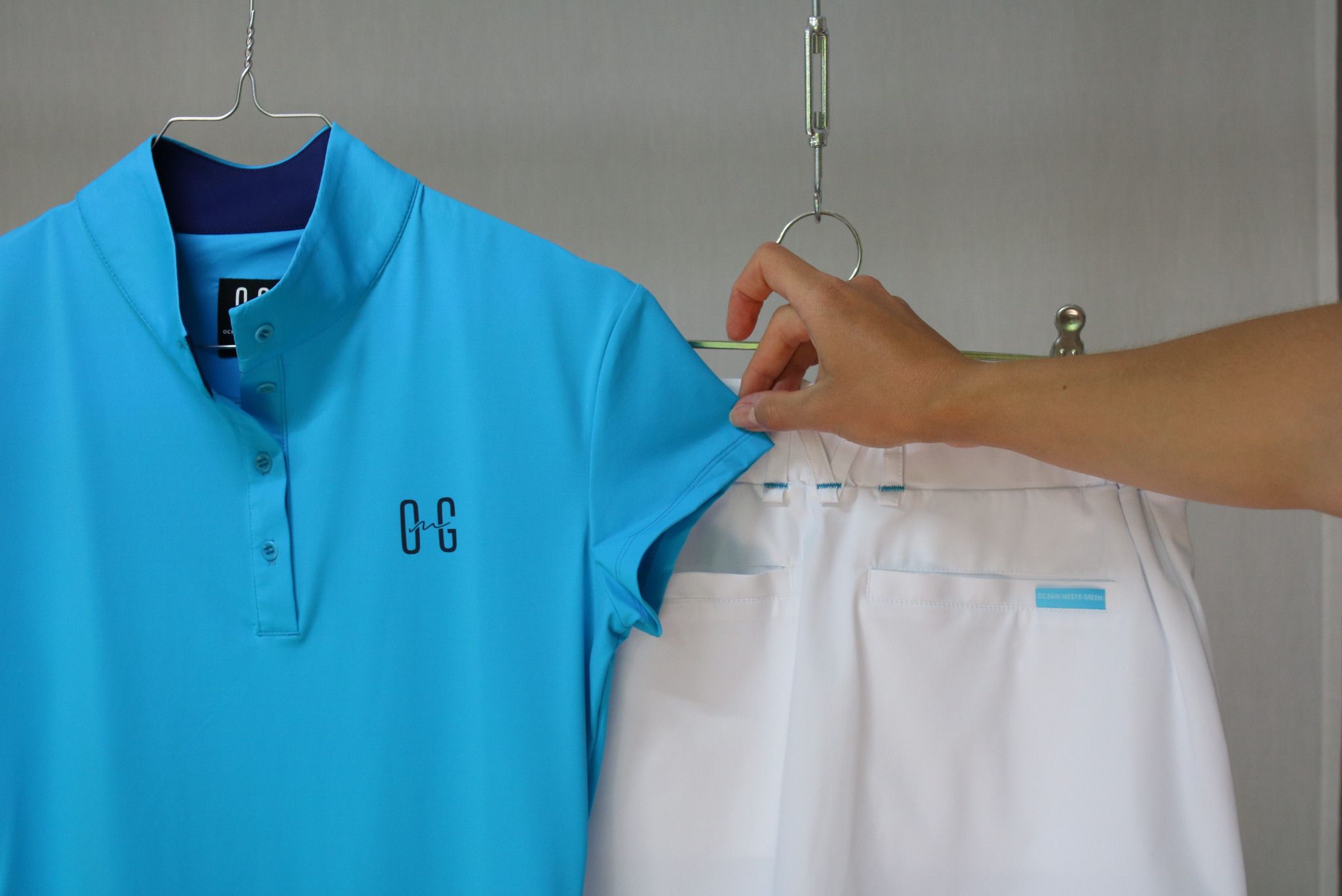 Turkusowa koszulka polo z ciemnoniebieskim kołnierzykiem i logo oraz białe spodnie na wieszakach, prezentowane przez osobę trzymającą koszulkę.