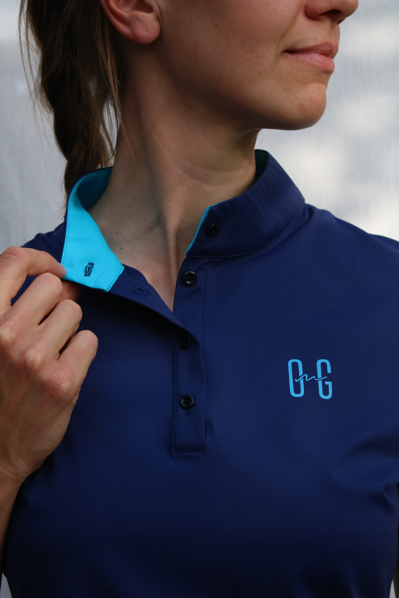 Kobieta w granatowej koszulce polo z turkusowym kołnierzykiem i wyhaftowanym logo, rozpinająca górny guzik.