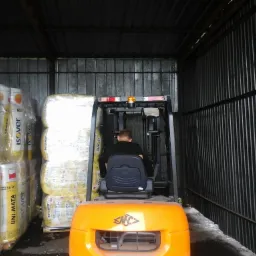 Wózek widłowy z operatorem transportuje paletę wełny mineralnej Isover w magazynie o konstrukcji stalowej z blachy falistej. Widok zza wózka.