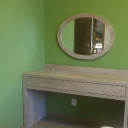 Toaletka z jasnego drewna z okrągłym lustrem na zielonej ścianie. Widoczne gniazdko elektryczne pod blatem.
