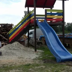Kolorowy plac zabaw z drewnianą konstrukcją, czerwoną dachówką i niebieską zjeżdżalnią, otoczony piaskiem i stertą drewna w tle.