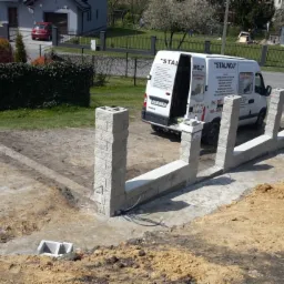 Wznoszenie muru oporowego z szarej kostki betonowej, widoczny zarys fundamentu, w tle biały bus z otwartymi drzwiami i logo firmy.