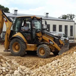 Żółta koparko-ładowarka CAT 432E podczas prac ziemnych na placu budowy, widoczne sterty ziemi i kamieni, w tle budynki i zaparkowany biały bus.