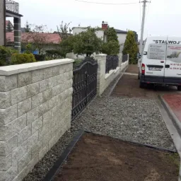 Nowo wybudowane ogrodzenie z szarej kostki łupanej z czarnymi metalowymi przęsłami, widok na świeżo przygotowany teren z wysypanym grysem i zaparkowany samochód dostawczy firmy budowlanej.