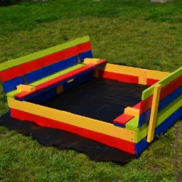 Kolorowa, drewniana piaskownica z siedziskiem, pomalowana w pasy: czerwony, żółty, niebieski i zielony, wyłożona czarną agrowłókniną, ustawiona na trawniku.