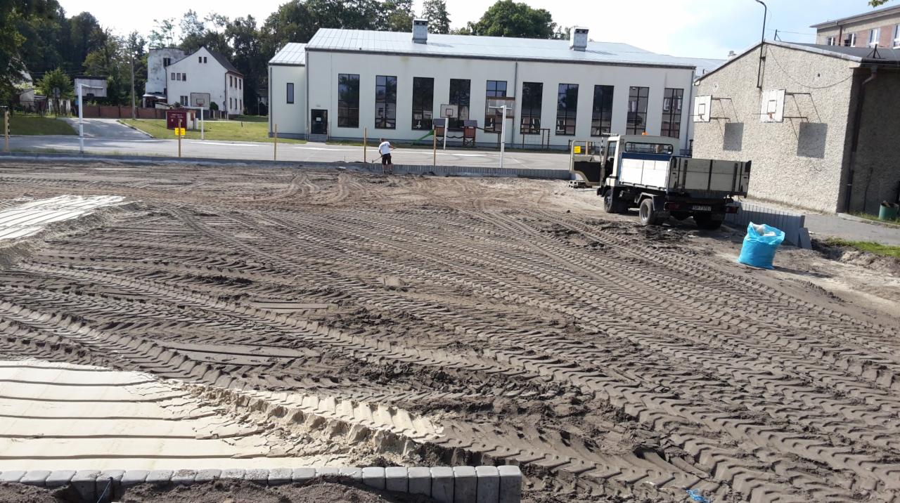 Teren przygotowany pod układanie nawierzchni z kostki brukowej, widoczne ślady opon ciężkiego sprzętu, częściowo ułożona podsypka i krawężniki, w tle budynek szkoły i zaparkowana ciężarówka.