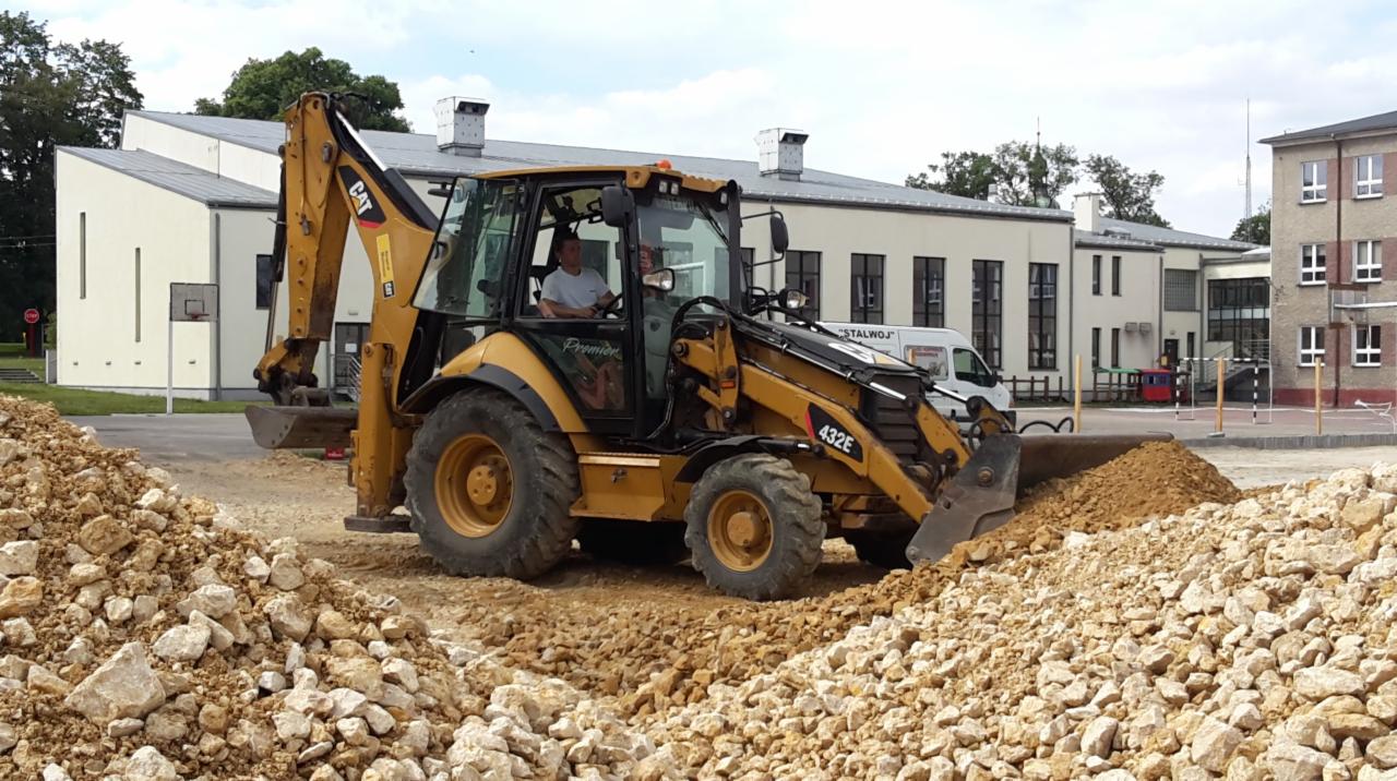 Żółta koparko-ładowarka CAT 432E podczas prac ziemnych na placu budowy, widoczne sterty ziemi i kamieni, w tle budynki i zaparkowany biały bus.