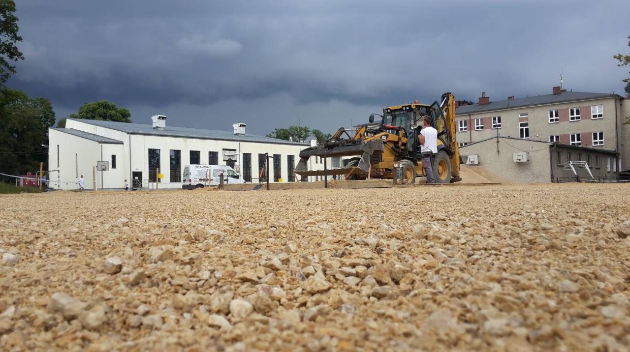 Żółta koparko-ładowarka wyrównuje teren pod budowę, widok z perspektywy gruntu na żwirową powierzchnię, w tle budynki i pochmurne niebo.