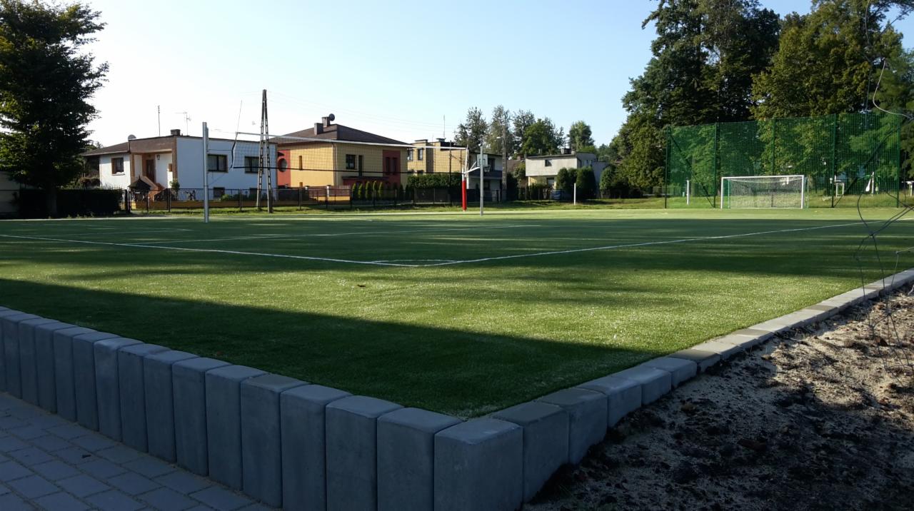 Nowo wybudowane boisko sportowe z zieloną sztuczną trawą, obrzeżem z szarych betonowych kostek i siatkami do siatkówki oraz bramkami do piłki nożnej w tle, otoczone drzewami i zabudową mieszkalną.
