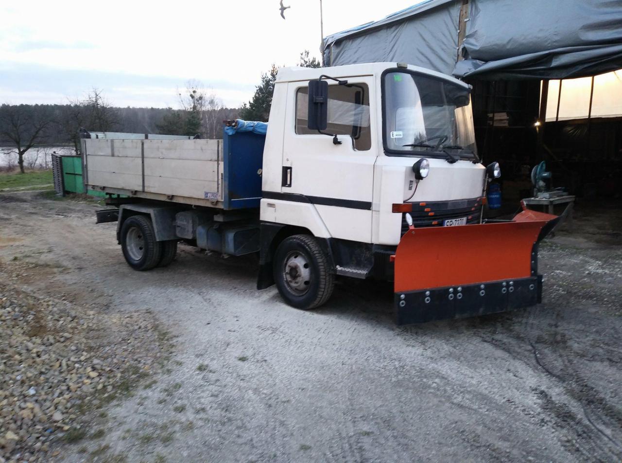 Biała ciężarówka z pomarańczowym pługiem do śniegu zaparkowana na żwirowym podłożu, widoczny fragment skrzyni ładunkowej i otoczenie z budynkami gospodarczymi w tle.