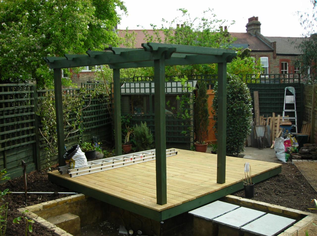 Konstrukcja drewnianego tarasu z zieloną pergolą w ogrodzie, drabina oparta o taras, widoczne narzędzia ogrodnicze i roślinność w tle.
