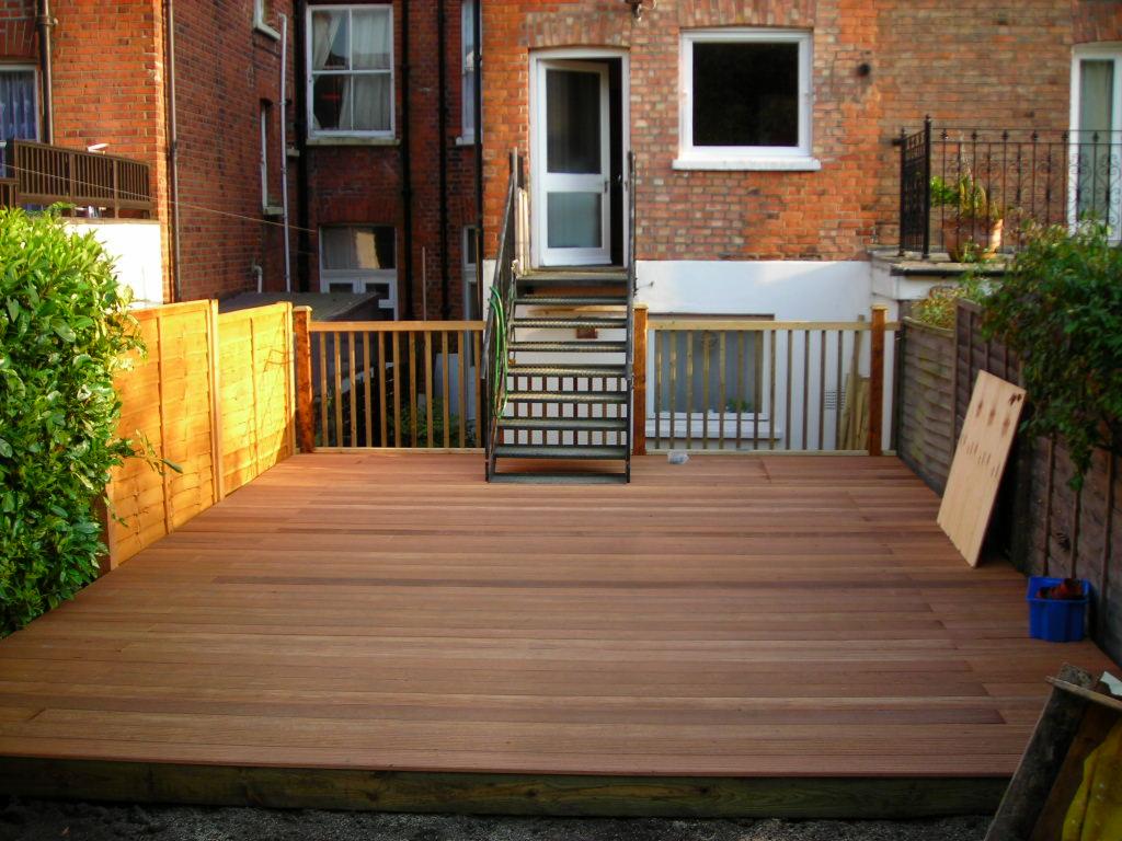 Nowoczesny taras z drewna egzotycznego, z widocznymi schodami metalowymi prowadzącymi do budynku z czerwonej cegły i fragmentami ogrodzenia drewnianego.