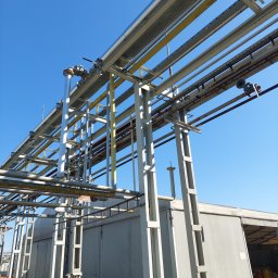 StalSpaw - Metalowa konstrukcja wsporcza z rurociągami i kablami na tle błękitnego nieba i szarego budynku przemysłowego.