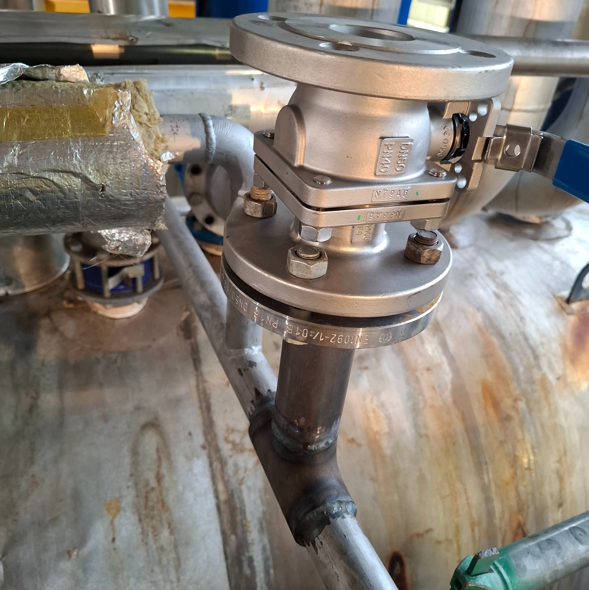 Metalowa armatura przemysłowa z zaworem kulowym, złączona śrubami i nakrętkami, z widocznymi spawami rur i izolacją termiczną na tle zbiornika przemysłowego.