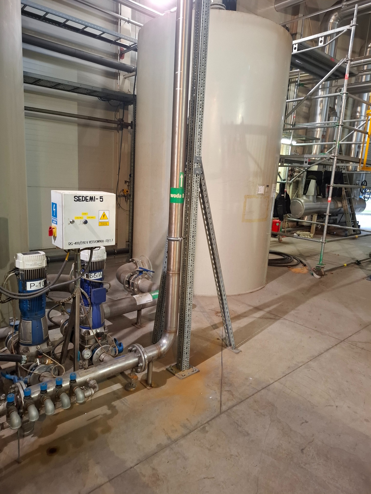 Instalacja przemysłowa z zbiornikami, rurami, pompami i skrzynką kontrolną Sedemi-5, widoczne rusztowania w tle.