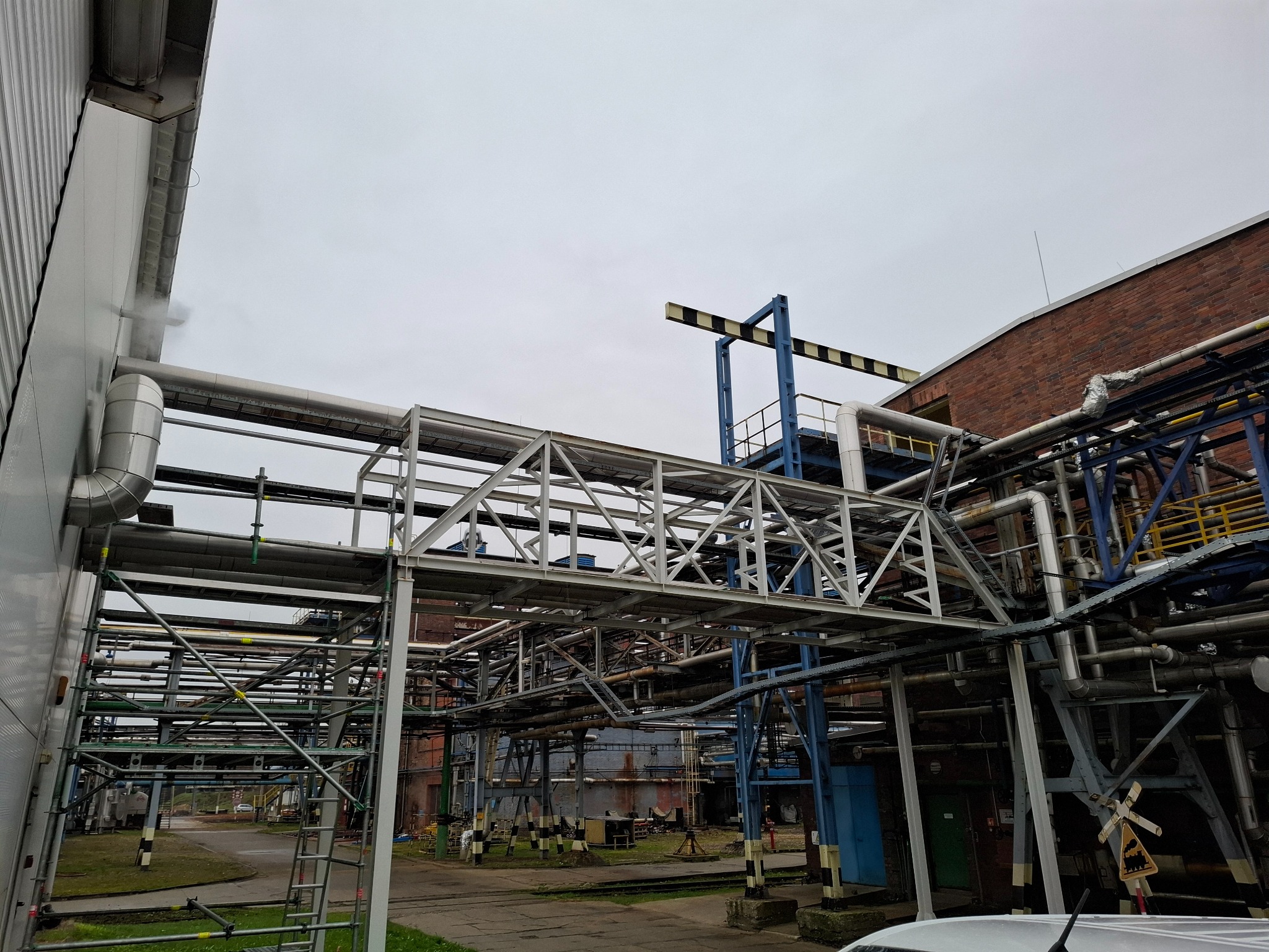 Skomplikowana instalacja rurociągów przemysłowych z metalowymi konstrukcjami wsporczymi, widoczne elementy rusztowań i znak ostrzegawczy na tle budynków fabrycznych.