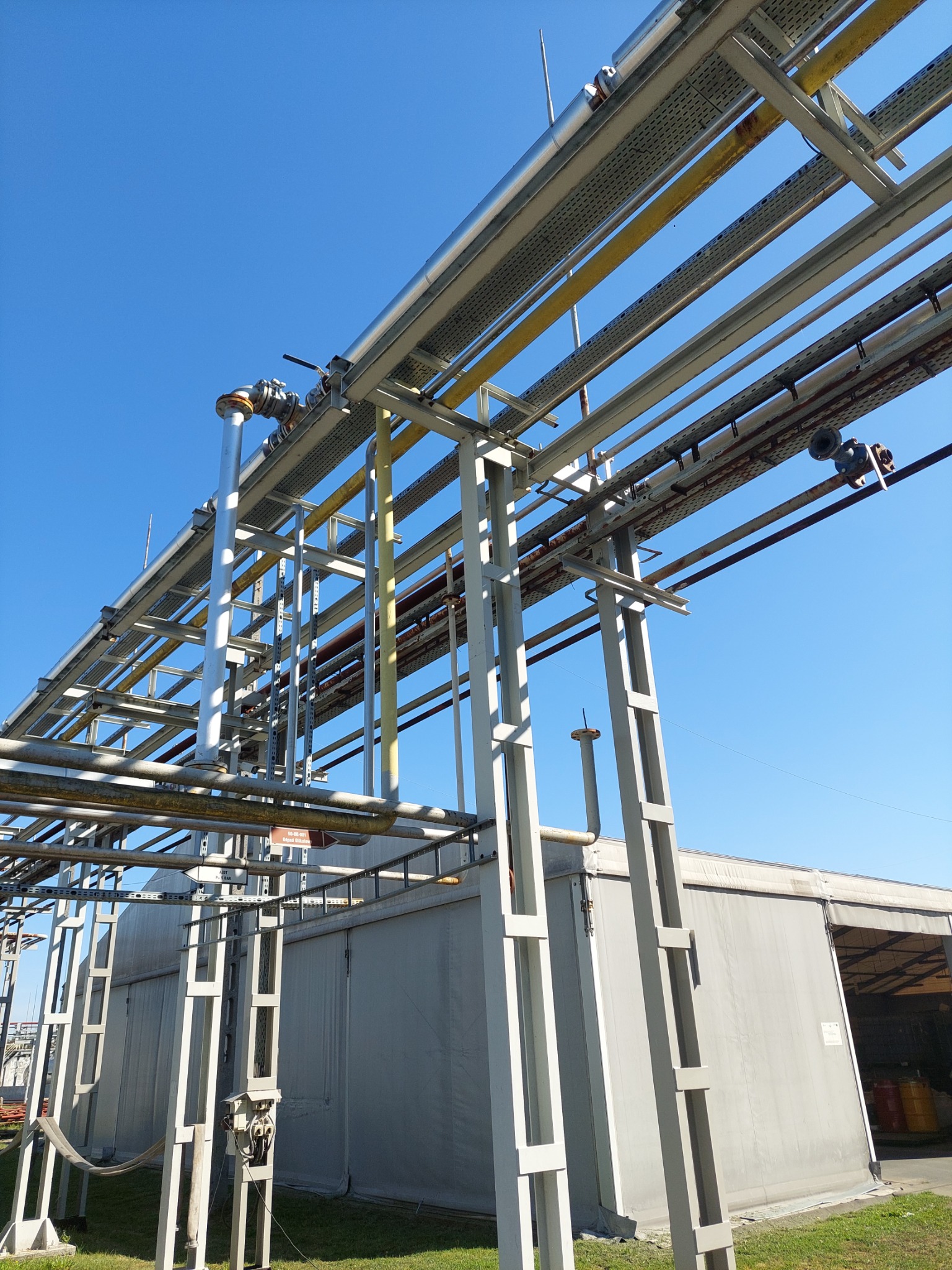 Metalowa konstrukcja wsporcza z rurociągami i kablami na tle błękitnego nieba i szarego budynku przemysłowego.