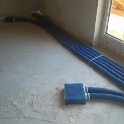 Niebieskie rury instalacyjne, częściowo ułożone w podłodze w surowym stanie, prowadzące do skrzynki rozdzielczej; widoczne fragmenty ściany i okna.