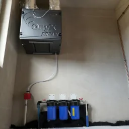 Szara centrala wentylacyjna Onyx Dream podwieszona pod sufitem, z podłączonymi przewodami wentylacyjnymi i zestawem trzech niebieskich filtrów wody na dole, w surowym pomieszczeniu.