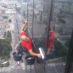 Mężczyzna myje okna wieżowca na wysokości z widokiem na Warszawę, używając profesjonalnego sprzętu i technik alpinistycznych.