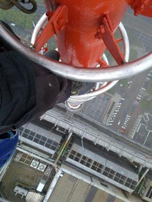 Wysokogórski widok z nogi pracownika na szczycie pomalowanej na czerwono konstrukcji, prawdopodobnie masztu, z widokiem na budynki w oddali.