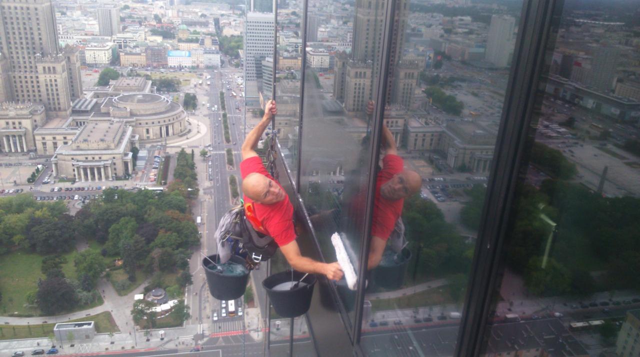 Mężczyzna myje okna wieżowca na wysokości z widokiem na Warszawę, używając profesjonalnego sprzętu i technik alpinistycznych.