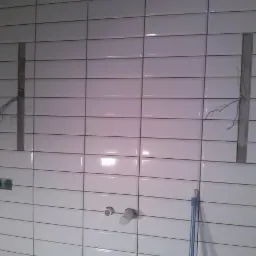Białe, prostokątne płytki na ścianie w trakcie montażu. Widoczne otwory na instalacje i wystające przewody. Częściowo ułożona glazura w łazience.