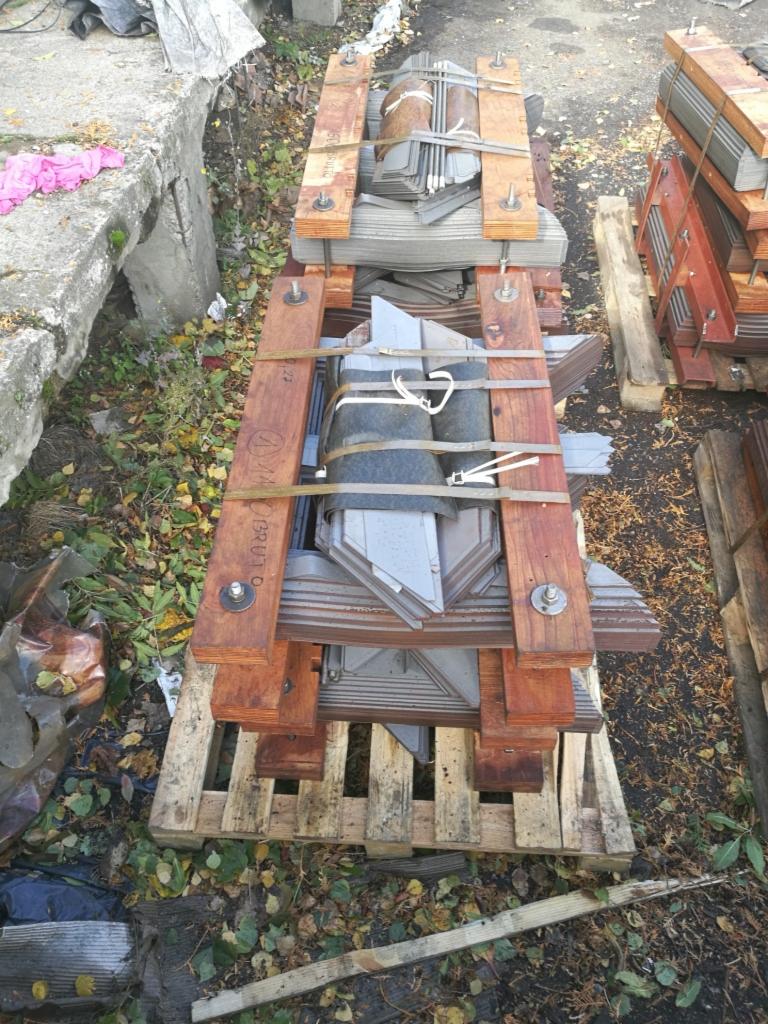 Stos metalowych elementów, prawdopodobnie blaszanych, zabezpieczonych drewnianą konstrukcją i taśmami, ułożony na palecie w otoczeniu liści i gruzu.