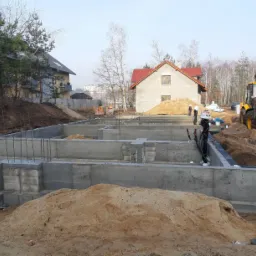 Wylane betonowe fundamenty domu jednorodzinnego z wystającymi prętami zbrojeniowymi, widoczny żółty traktor i koparki ziemi w tle, dwóch pracowników w białych kaskach oraz częściowo wybudowany dom...