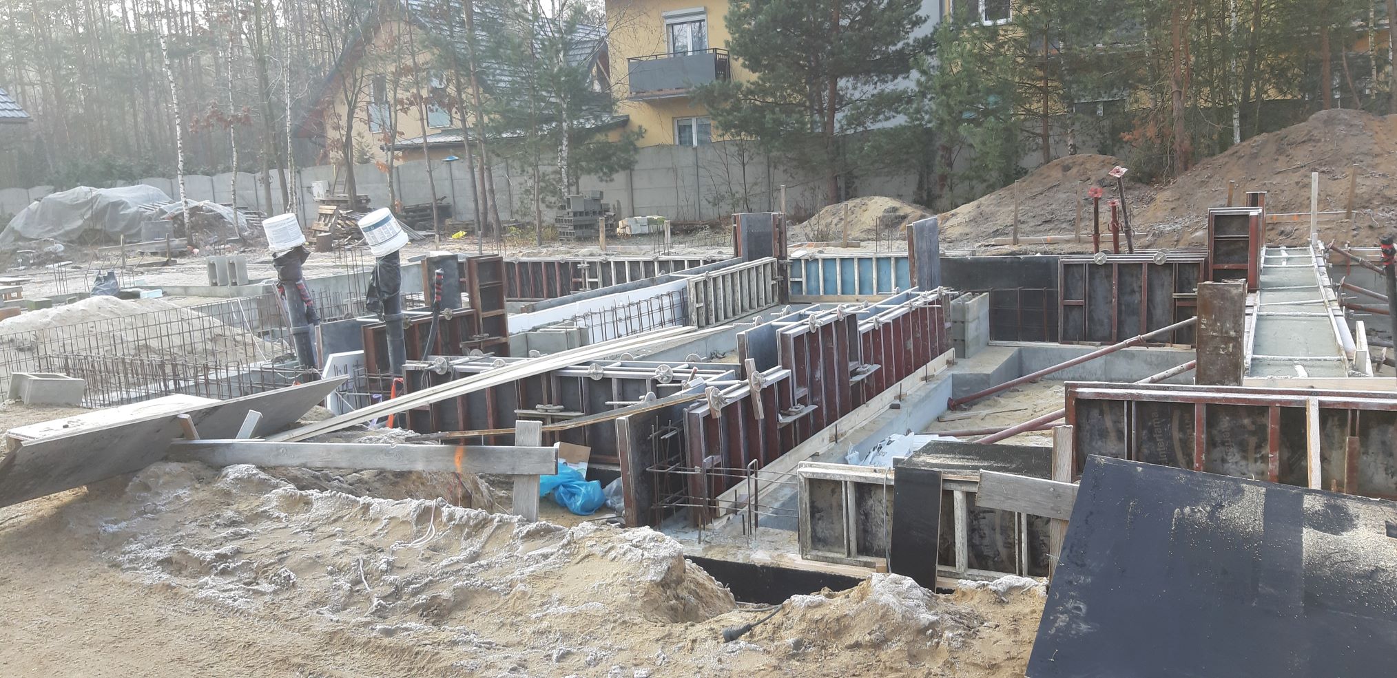 Budowa fundamentów domu jednorodzinnego, widoczne szalunki zalane betonem, zbrojenie, teren budowy w zimowej scenerii, w tle dom w trakcie budowy i drzewa.