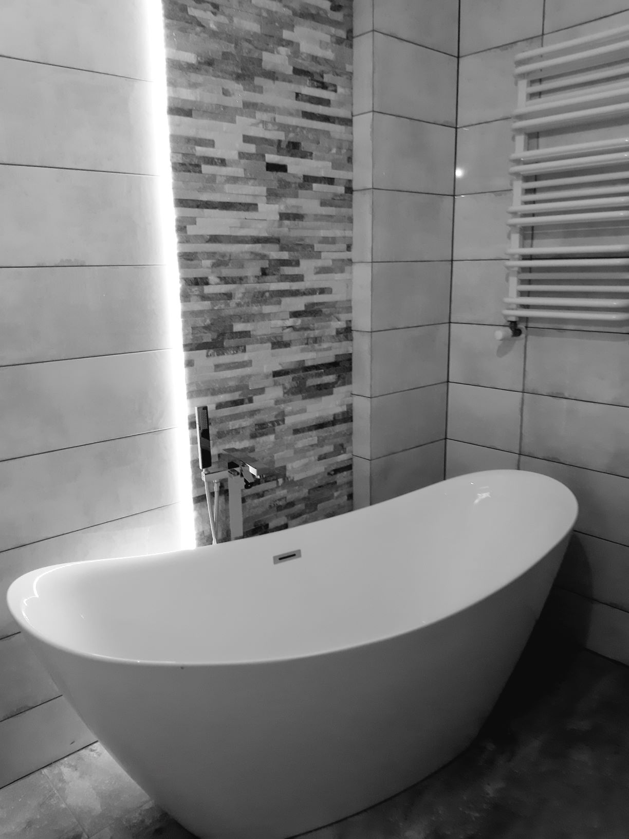 Nowoczesna łazienka w odcieniach szarości z wolnostojącą wanną, mozaiką na ścianie podświetloną listwą LED i grzejnikiem drabinkowym.