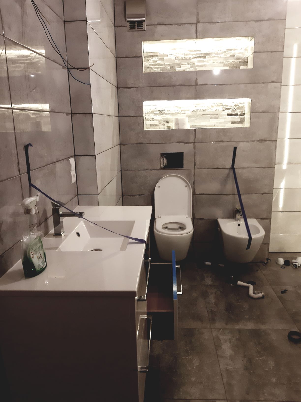 Łazienka w trakcie wykańczania: widoczna umywalka z szafką z otwartą szufladą, toaleta, bidet, płytki na ścianach oraz elementy instalacji hydraulicznej na podłodze.