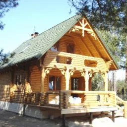 Jasny dom z bali z zielonym dachem i zdobieniami z drewna, widoczny z przodu, z fragmentem przyczepy kempingowej w tle. Słoneczny dzień.