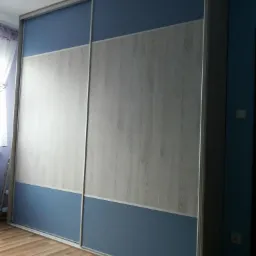 Dwudrzwiowa szafa przesuwna z niebieskimi i drewnopodobnymi panelami, srebrne obramowanie, widoczna podłoga z paneli i fragment lampy sufitowej.