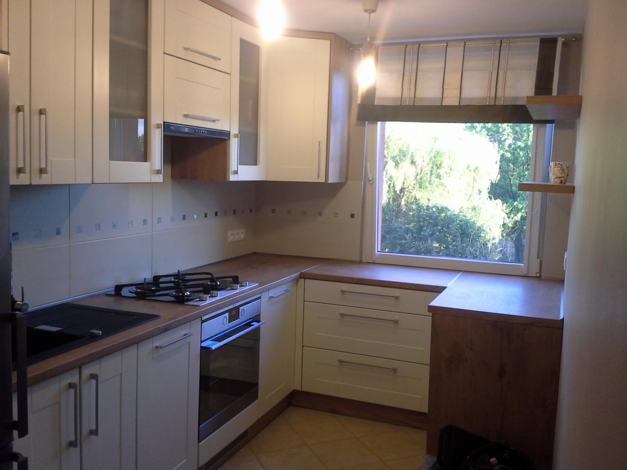 Jasna kuchnia z drewnianym blatem, okno z widokiem na zieleń. Płyta gazowa i piekarnik w zabudowie. Górne szafki z przeszkleniami.