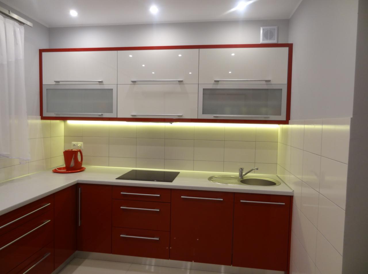 Nowoczesna kuchnia: biało-czerwone szafki z podświetleniem LED, szklane fronty górnych szafek, ceramiczny blat i płytki. Widoczny zlew i płyta indukcyjna.